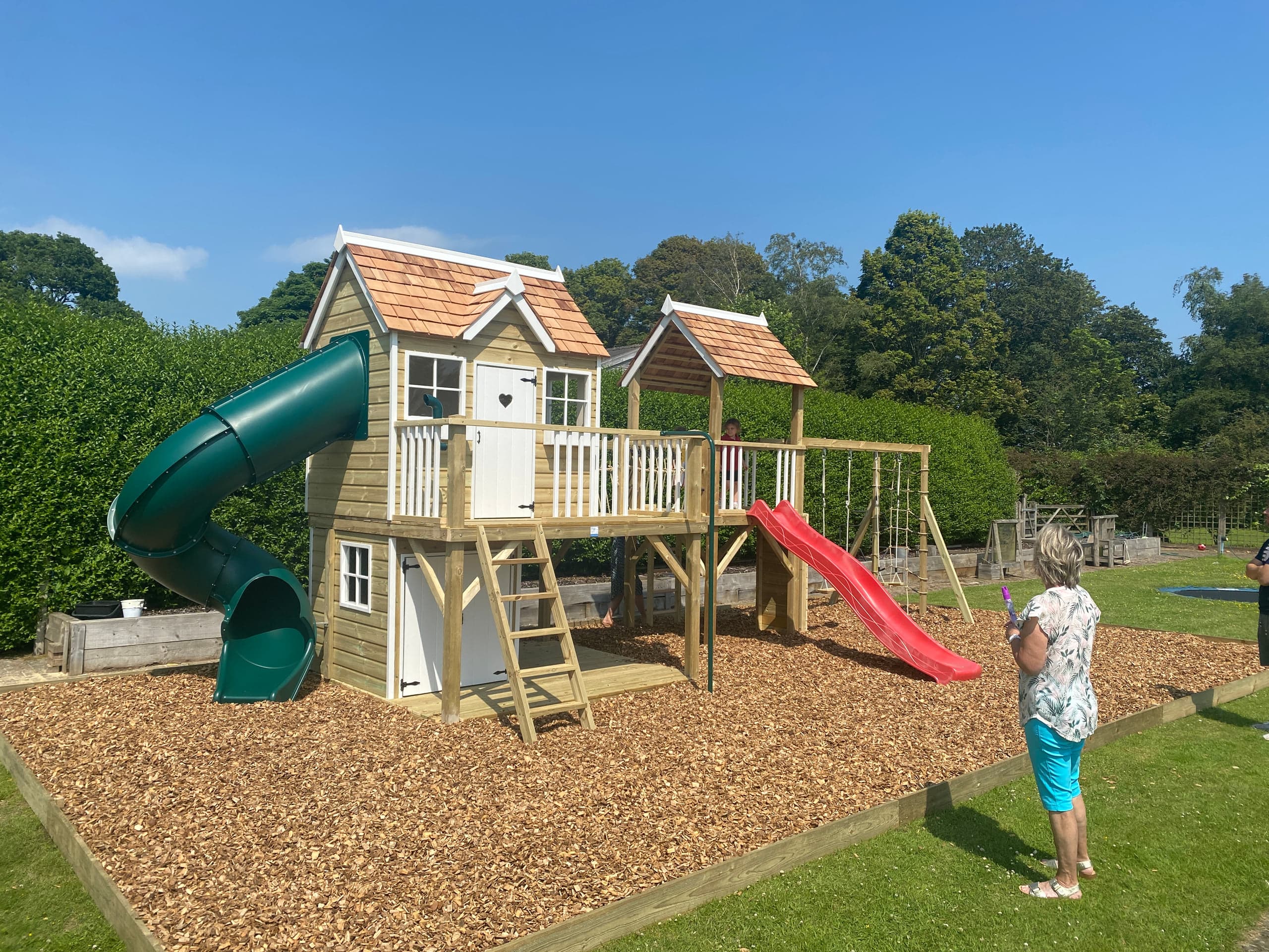 A cedar-shingled playhouse with a green tube slide and a red wave slide, an adult and child at the play deck