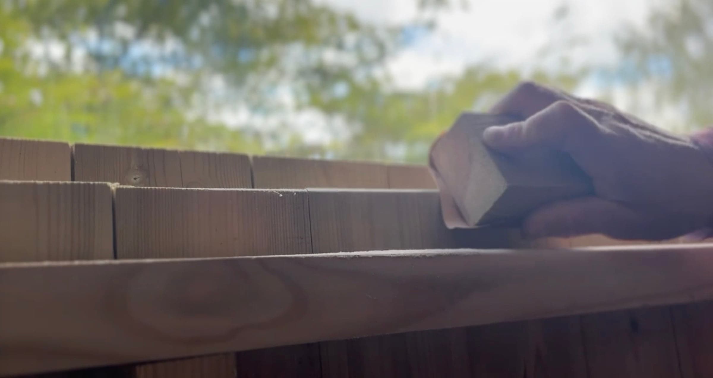 A hand sanding shingle courses at the workshop window, garden light beyond